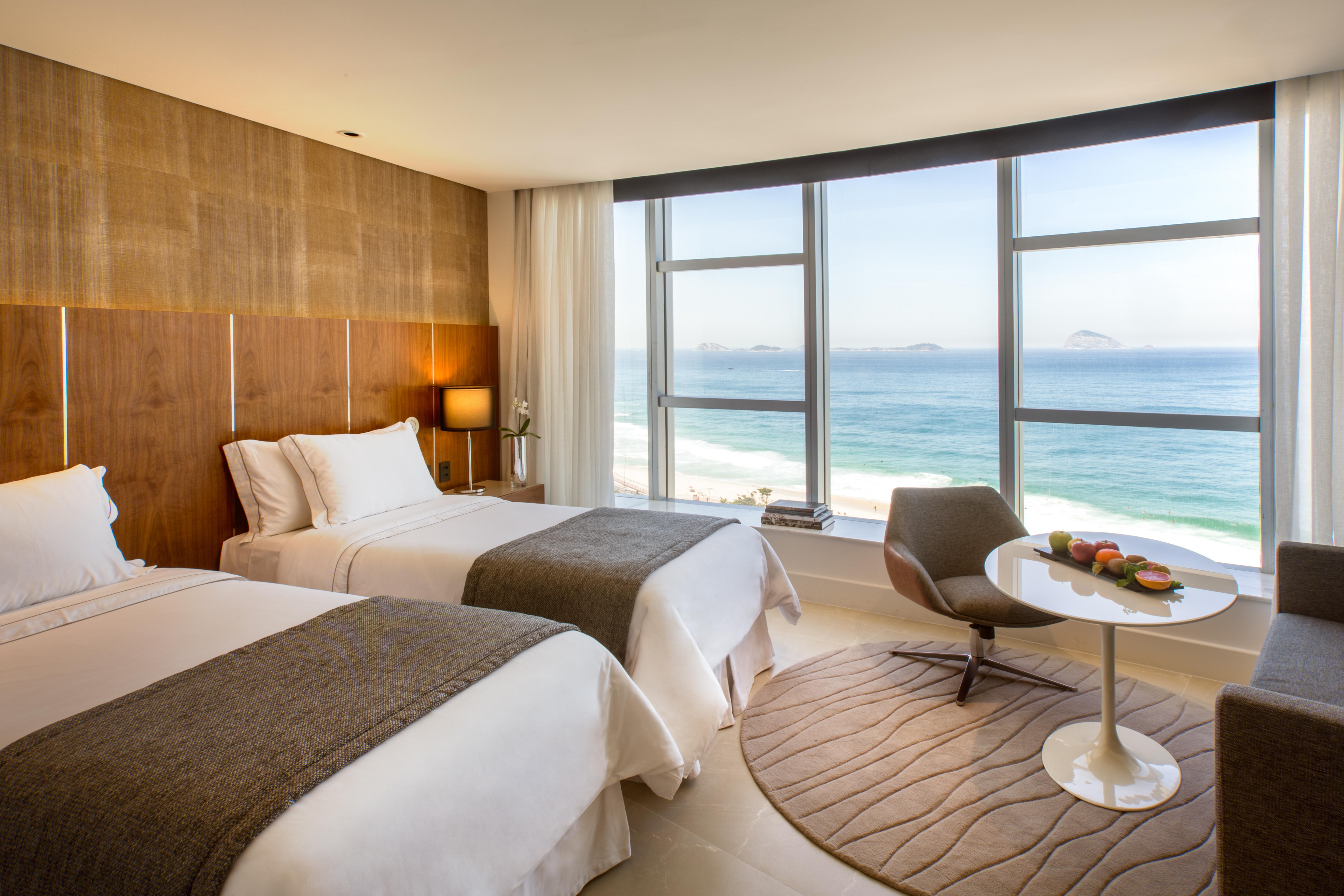 Hotel Nacional Rio De Janeiro - Oficial Exteriér fotografie The photo shows a modern hotel room with two beds adorned in white linens and gray throws. Large windows allow natural light to flood the space, offering a stunning view of the ocean. In the corner, there's a comfortable chair and a small round table